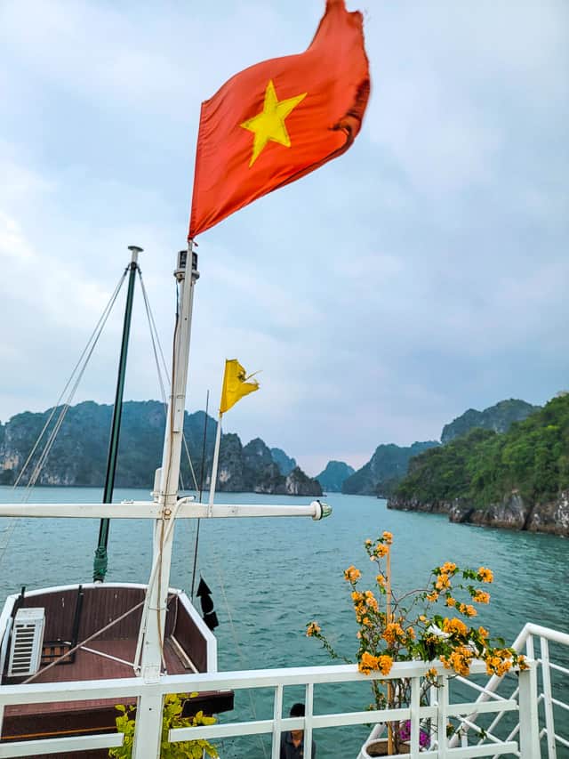 The front of a ship with the Vietnamese flag flying (red flag with yellow star) is in the foreground and halong bay is in the background.