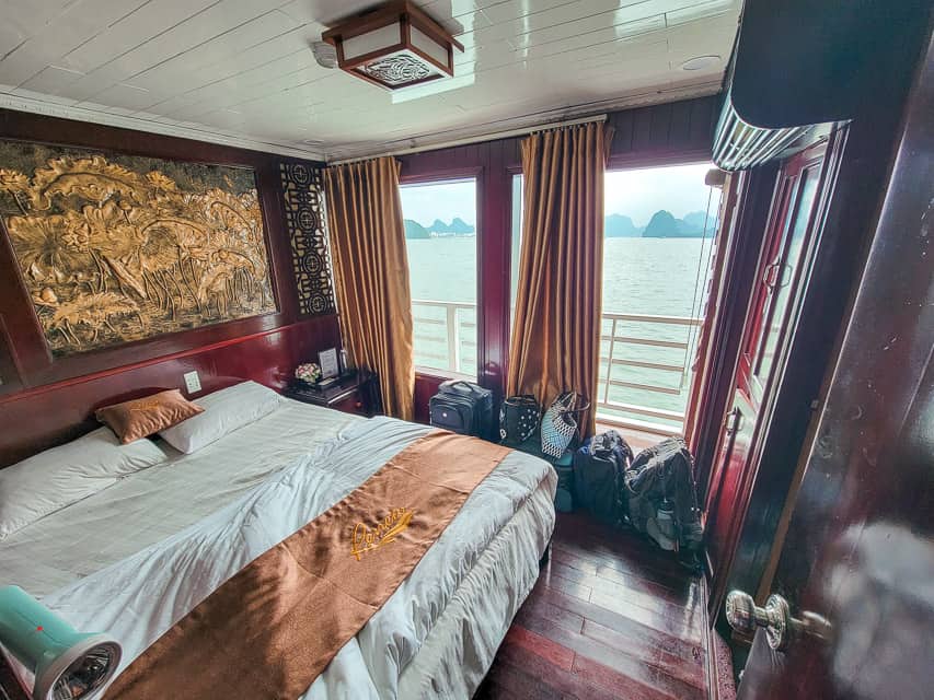 The room along a Halong Bay cruise with a king size bed with white sheets and a deck that shows view of the teal a water and floating islands.