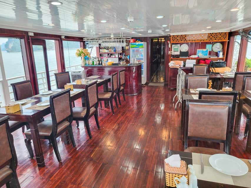 The dining room of the Halong Bay cruise. There are several tables set for 4 set up, deep wooden floors, a bar in the background, and floor to ceiling windwos.