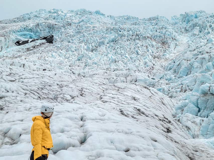 A woman stands on the base of a tall ice hill with craggles of ice formations on the side of the glacier.