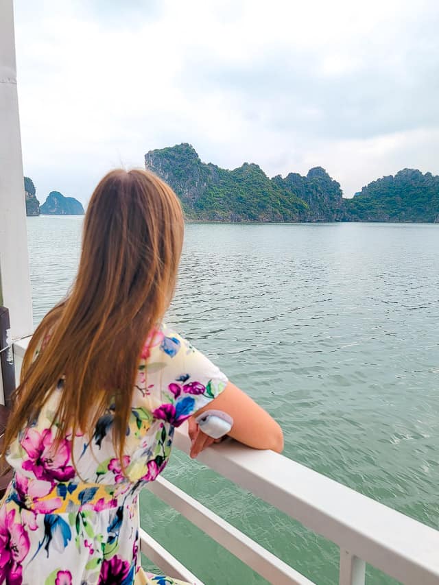 A woman in a flower dress sits on a balcony and watches the scenery pass in this Halong Bay alternative cruise.