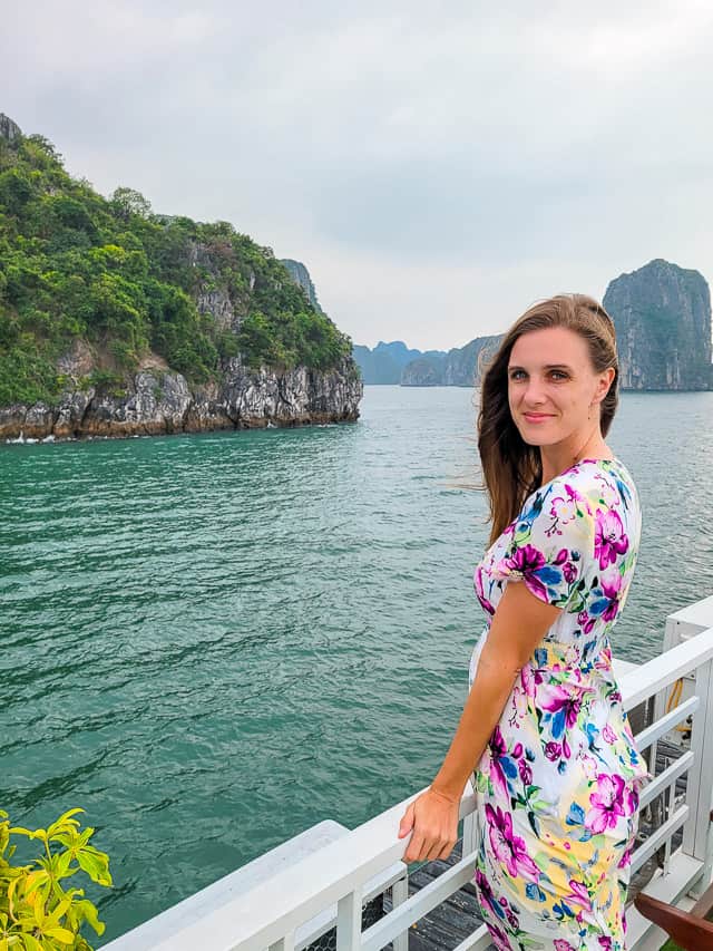 A woman in a white floral dress leans on the outside railing of the cruise looking at the rock islands in Halong bay with lots of ferns and overbrush covering the islands.