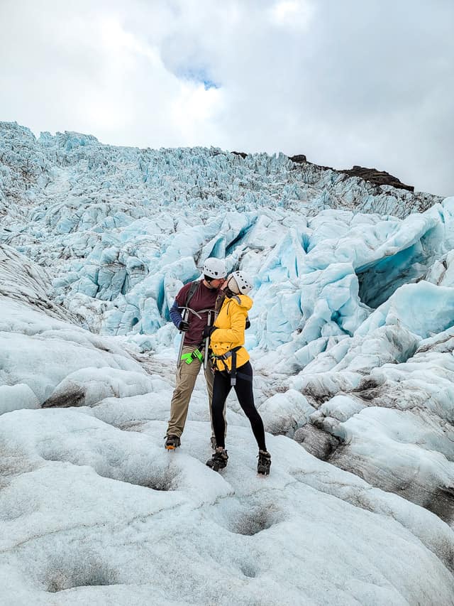 A man and woman in climbing gear, kiss on the base of a blue glacier.