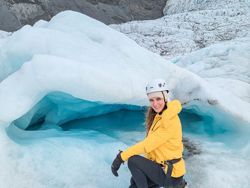 A woman squats by a small cavern of a melting snow cave with deep blue water.