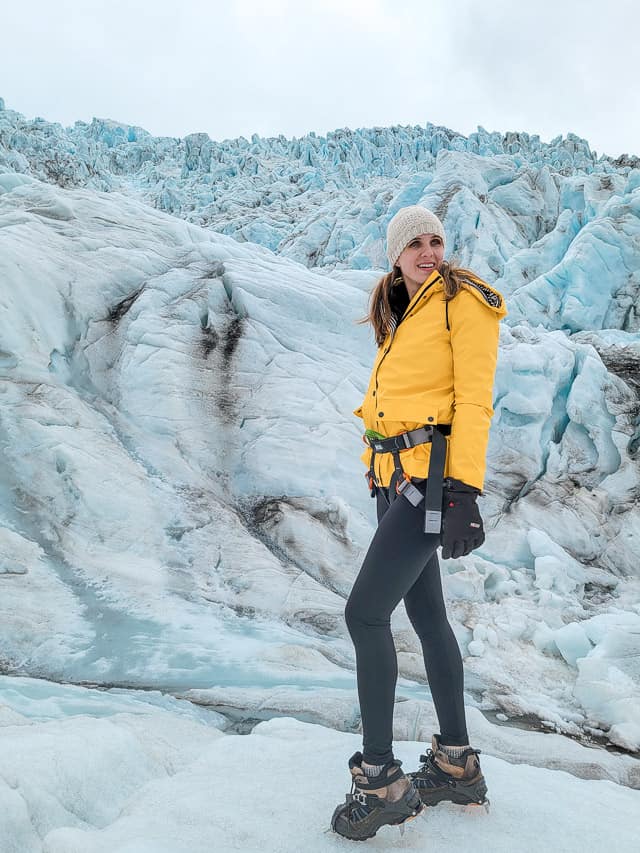 A woman stand in a yellow puffy jacket on the base of a gorgeous blue glacier.
