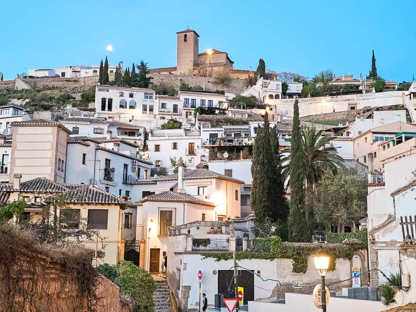 From a lower vantage point, looking up a hill at the white houses in the Albaicin neighborhood. Houses have clay roofs, ther are several palm and cypress trees dotted throughout, and the tower of a church is at the top of the hill.