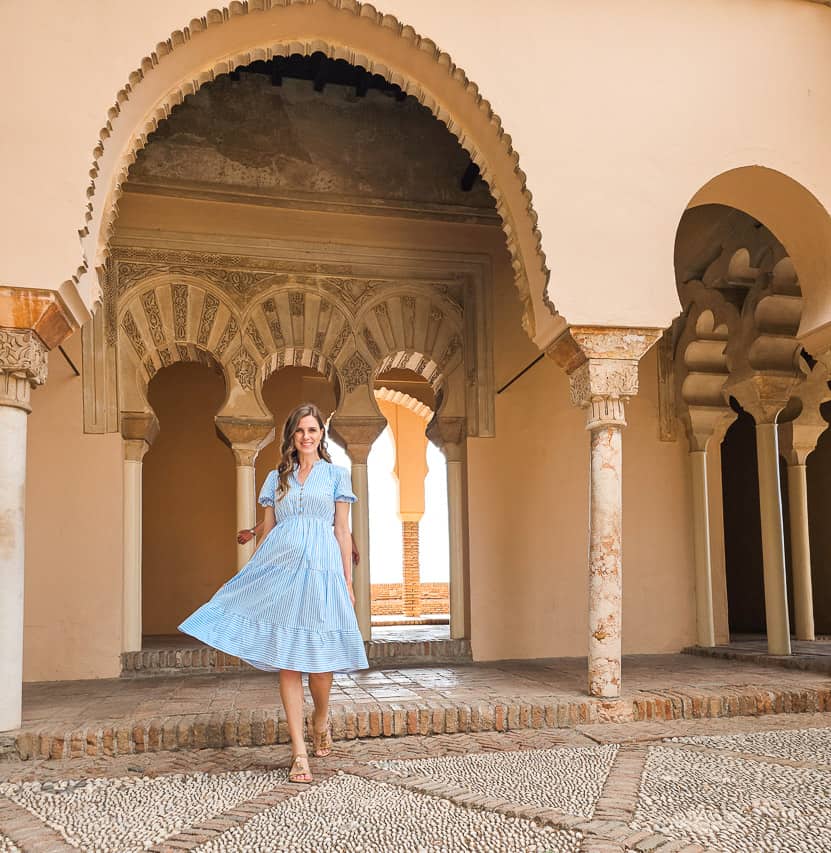 A woman in a blue and white striped dress smiles at the camera as she walks bewteen Islamic arches in a large courtyard.