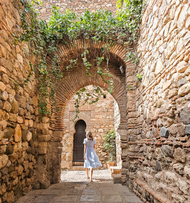 A woman in a blue and white dress walks away from the camera, down a cobblestoned passageway with a large arch at the end and ivy hanging down. The Alcazaba is one of the top reasons to visit Malaga.