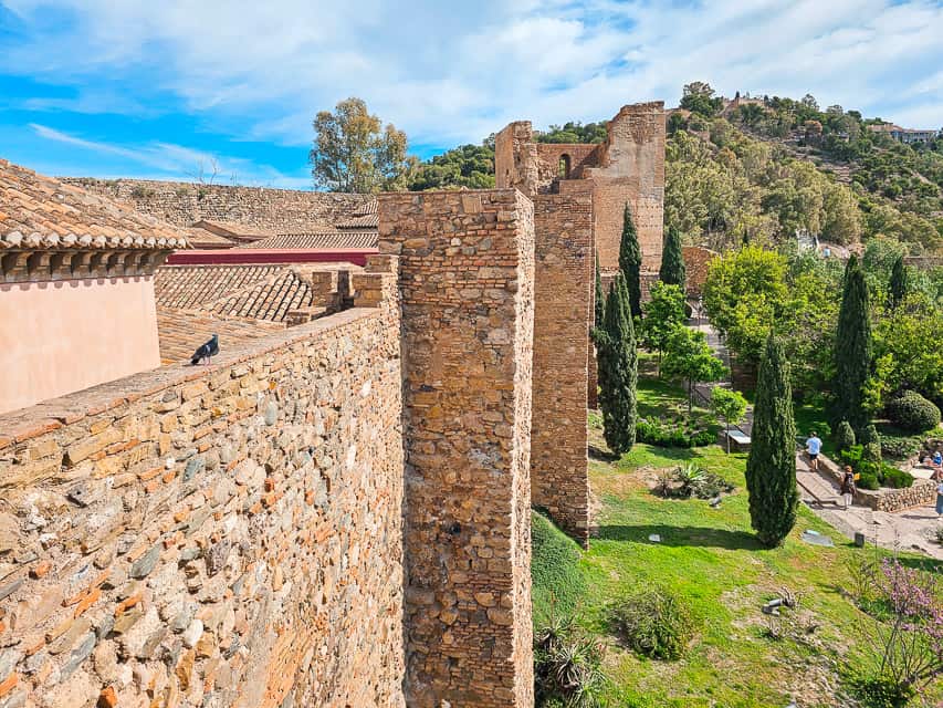 The tall walls of the Alcazaba, with towers spaced regularly, and green countryside below and around it.