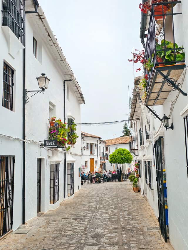 A cobblestone street runs between a whitewashed building leading to a cafe with many people dining.