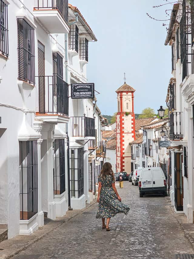 A woman walks down a little street in Grazalema - with white buildings with wrought iron black windows, and the red and white tower of a church is framed perfectly between the streets.