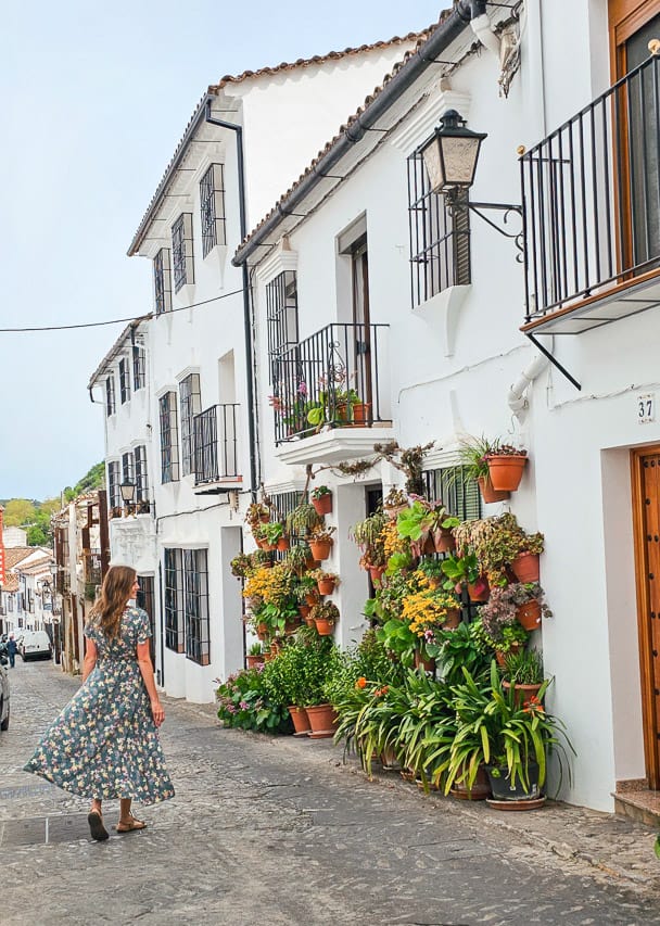 A woman walks down a street admiring a vibrant display of plants in Grazalema, Spain.