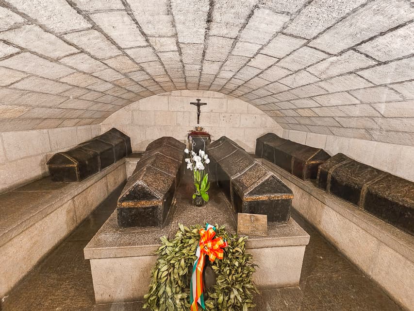 5 old, black coffins sit on low stone tables in an underground vault with a curved ceiling.