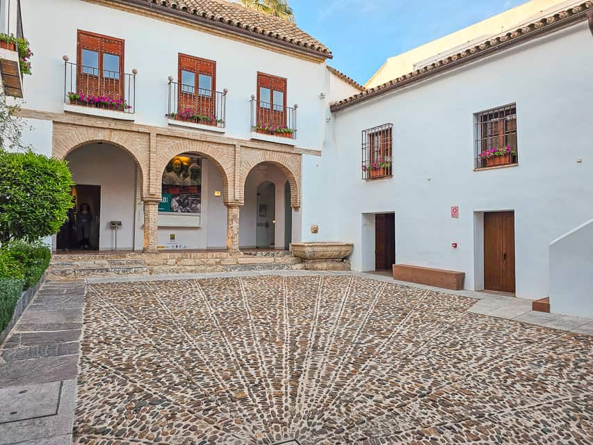 A patio with a sunburst shape in the cocbblestoned floors, a brick archway in the back, and white walls of the patio. There is just one bush on the left side of the patio.