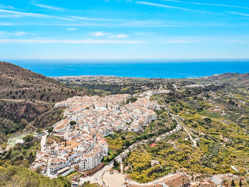 The view from the top of a large hill looking center down at the white buildings at the city of the Frigiliana at the edge of the coast of the blue sparkling ocean.