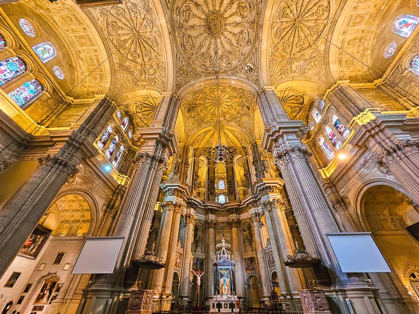The interior of the cathedral, with tall, wide columns, soaring arched ceilings, stained glass just under the roof, and intricate carvings on the ceiling itself. The cathedral is one of the reasons to visit Malaga.
