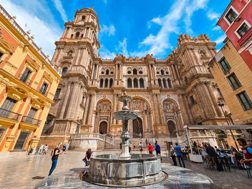 The tall, imposing cathedral of Malaga is the focal point, with only the left bell tower built. There is a fountain in the foreground, and buildings to the side of the cathedral. A few people mill about.