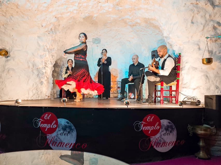 A woman in a black top and red skirt, dances flamenco dances in a cave with white walls. 2 woman and 2 mend are in the background, either singing, clapping, or playing the guitar.