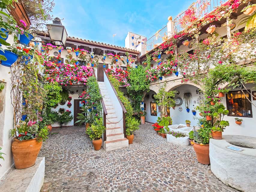 This small, private patio has cobblestoned flooring, a little well off to the side, and two stories that are covered in greenery, colorful pink, purple, and red flowers, and has a little staircase down the center.