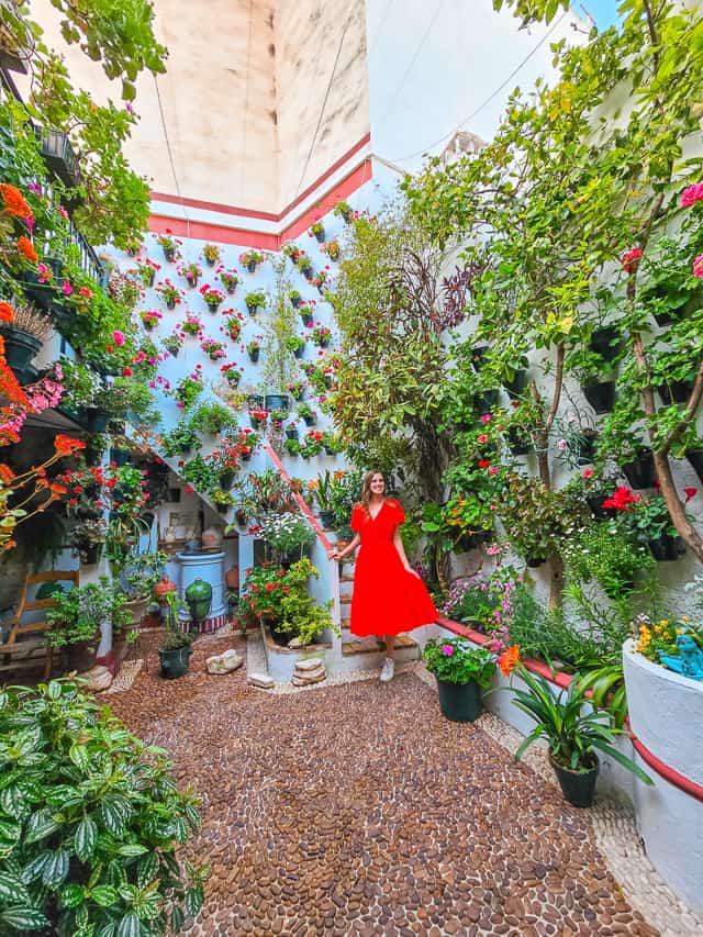 A woman in a red dress walks down a staircase at the back of a courtyard in Cordoba, that has white walls, cobblestoned flooring, plants and trees and flowerpots all over the walls.