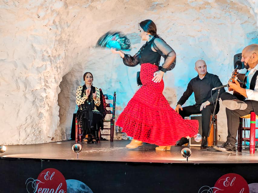 A woman in a black top and red skirt, holding a fan, dances flamenco dances in a cave with white walls. A woman and 2 mend sit on chairs in the background, either singing, clapping, or playing the guitar.