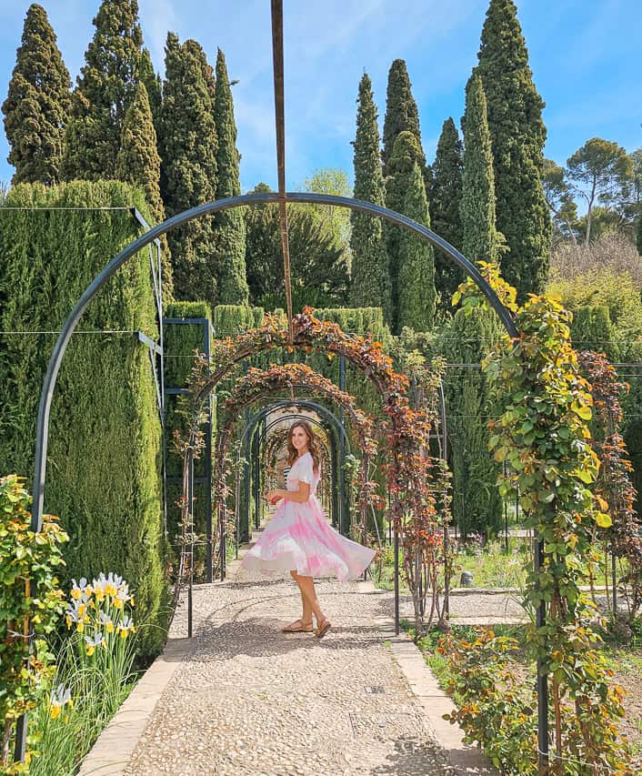 A woman in a pink and white dress twirls in the middle of the tall hedges of the Generalife gardens in Alhambra. The beautiful gardens here are on the reasons that Granada is worth visiting.