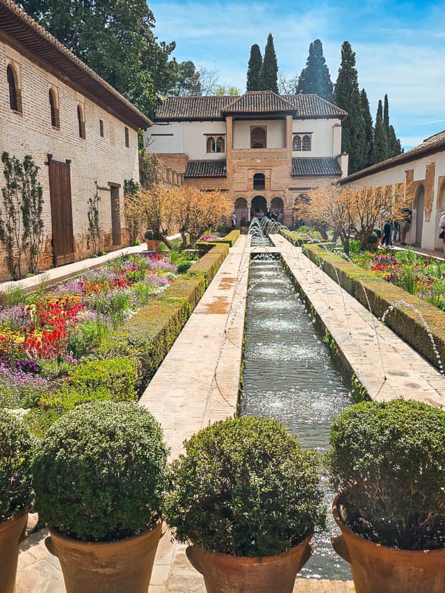 The Patio of the Acequia in Generalife in Alhambra. A long, skinny garden is surrounded by a buillding and a walkway, with a long basin of water in the middle with little fountain streams going in, and manicured flower gardens on each side of the basin.