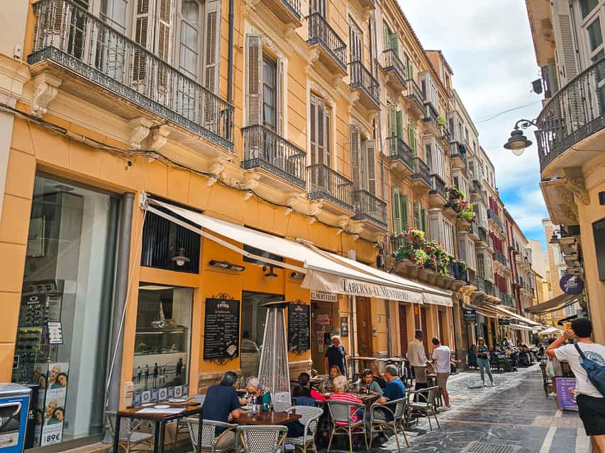 A typical old town street in Malaga, with tan walls, a restaurant with a patio on the street, lots of full length windows, and little balconies.