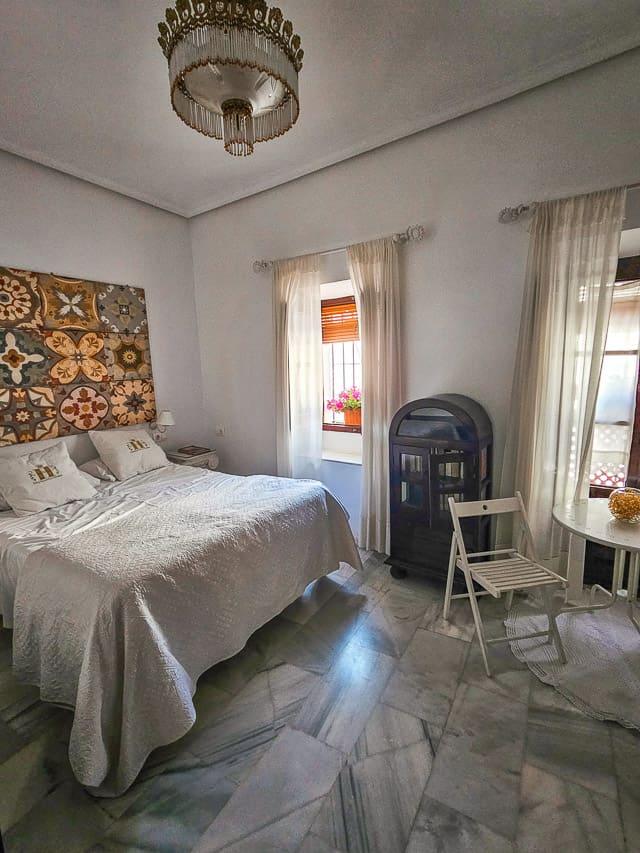 A hotel room with a double bed, marble flooring, an old chandelier, a little table and chair int he corner, and two windows with shuttlers and curtains.