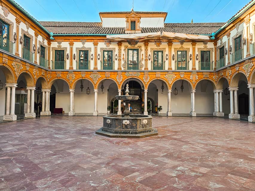 A rather large courtyard with pink and white marble tiles, a black marble fountain in the middle, orange arches on top of white columns, with green windows on the second level of the building.