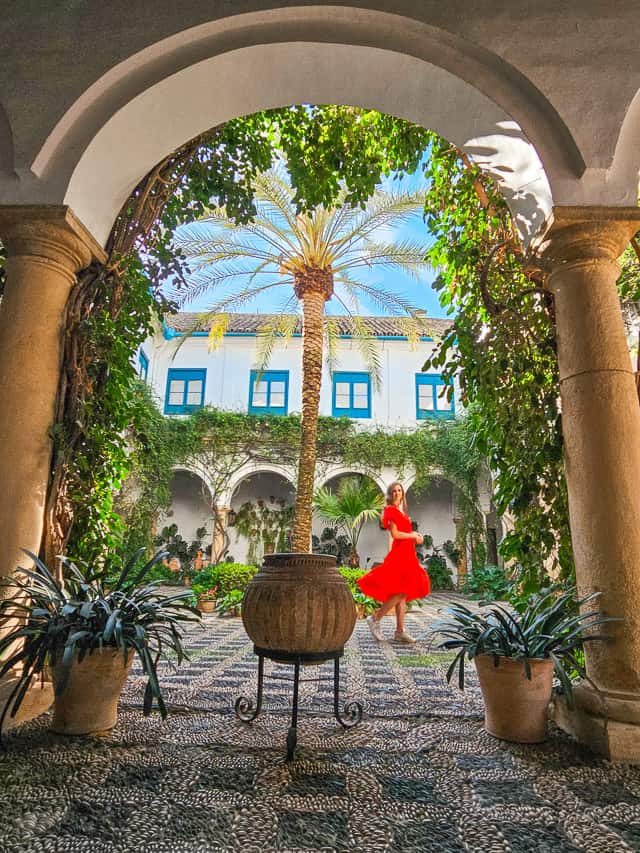 A girl in a red dress stands behind arches, in a courtyard with tiles and greenery.