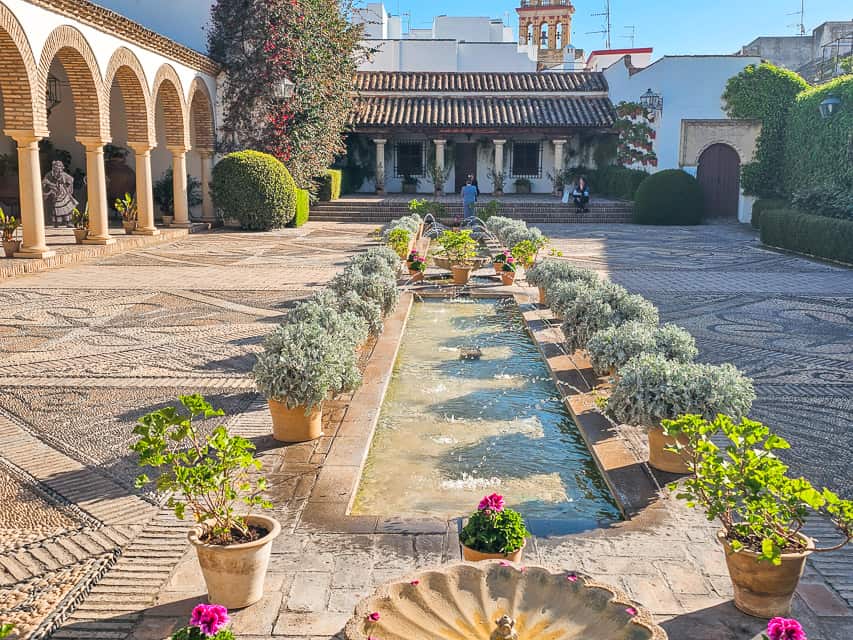 A rather large courtyard has an archway on the left side, vairous designs in the floor, a rectangular water basin in the center, with small jets of water sprinkling into the pond. In the background is a small portico with steps leading up to it.
