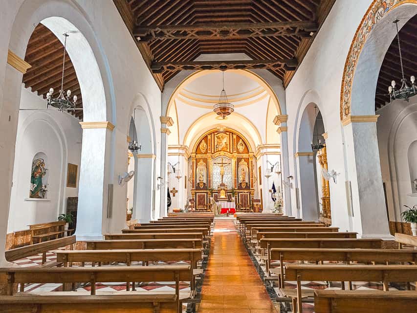 A church with multiple wooden pews with white pillar arches with a golden cross and paintings at the front of the room.