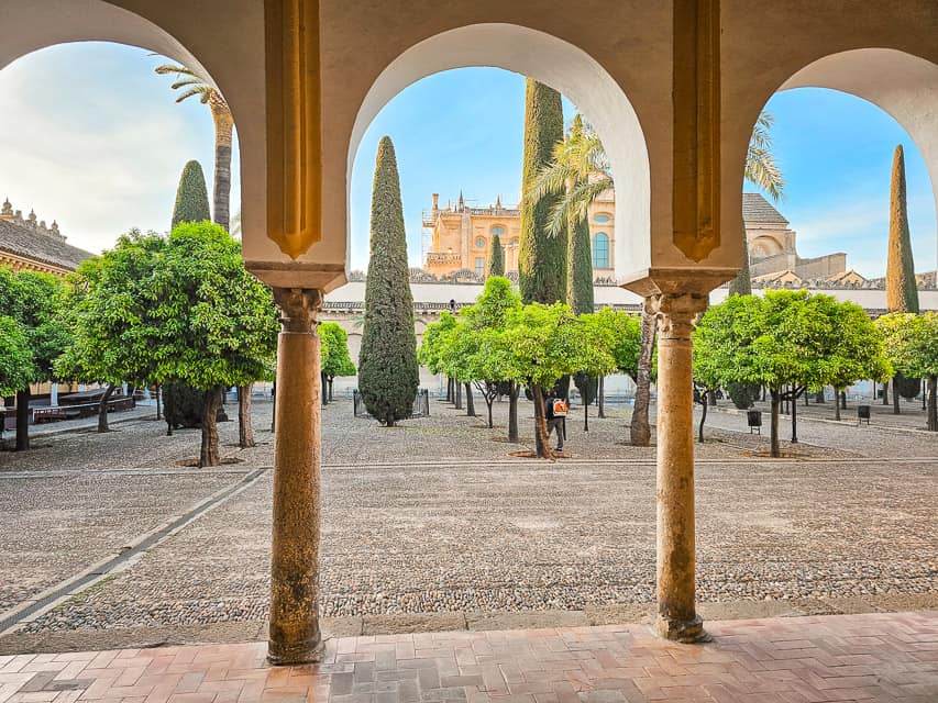 An archway with 3 arches is in the foreground, and a large courtyard filled with symmetrical lines of orange trees is in the background. Tall, skinny cypress trees go down the middle of the patio.