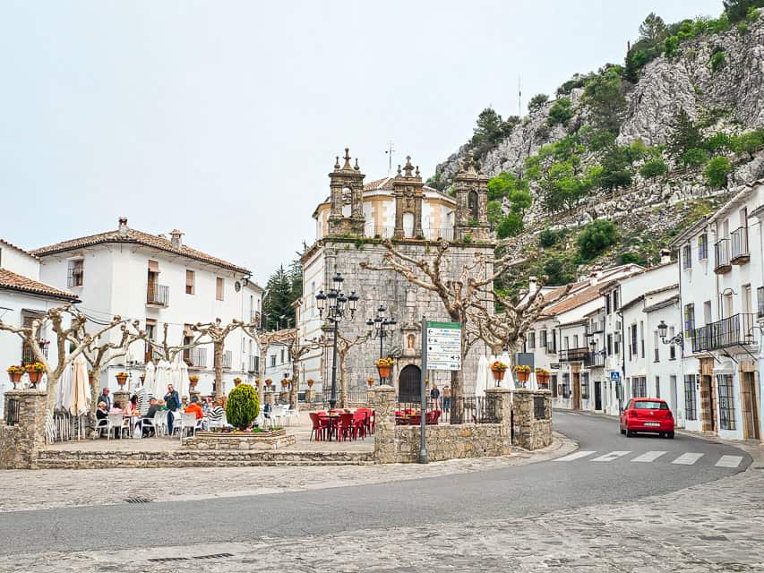 A view of the town square in Grazaelma - a cobblestoned square with a very short wall, a historic stone church on one end, a street going by another end, and white houses lining the square.