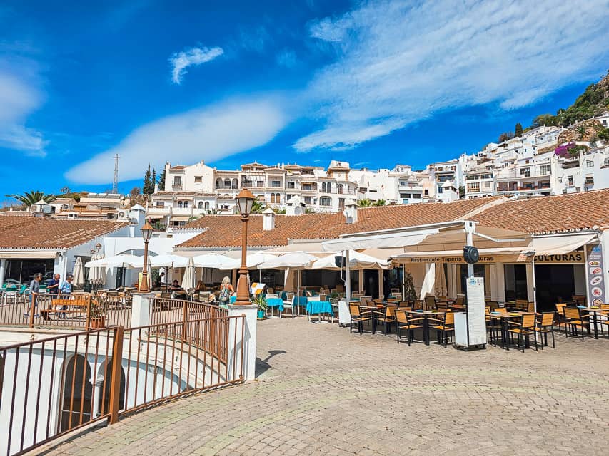 A cute restaurant with white and brown overhangs and multiple brown tables and chairs by the edge of the wall looking at the buildings above.