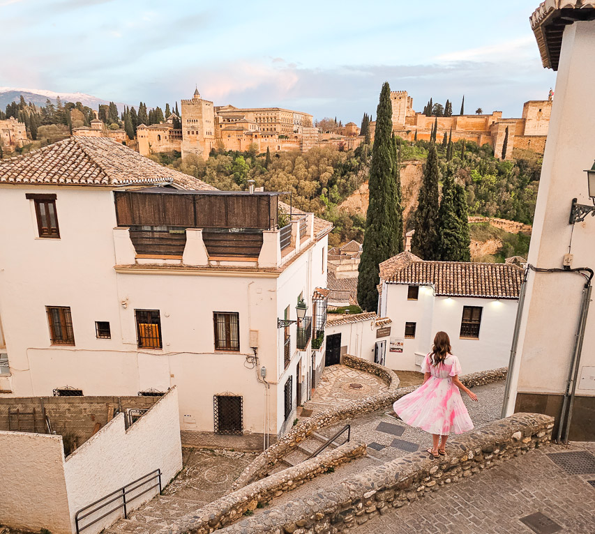 A woman in a pink and white dress stands among the white houses of the historic Albaicin neighborhood during the golden hour just before sunset. The grand structure of Alhambra can be seen on a hill in the background. Enjoying the viewpoints is a great reason to visit Granada.