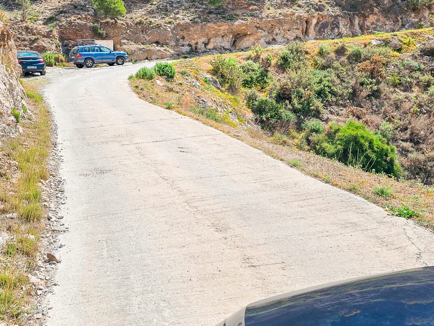A cement street winding up the rocky hill with 2 blue cars on the side of the road.