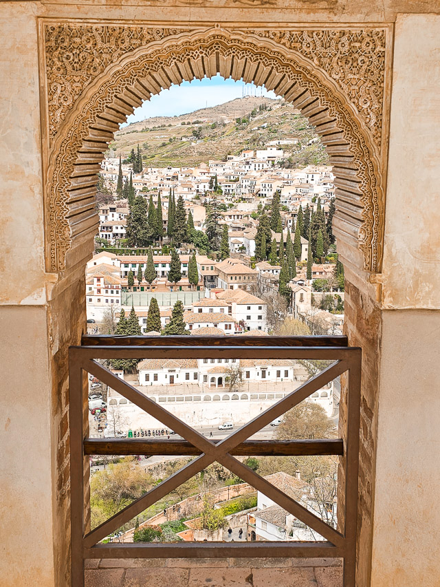 A view to the white houses and cypress trees of the Albaicin neighborhood through a fancy window opening in Alhambra.