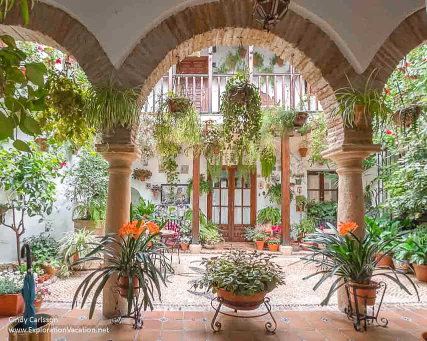 This picture looks through the stone and bricked archway into a small, private patio, that has beautiful wood accents and hanging greenery pots everywhere.