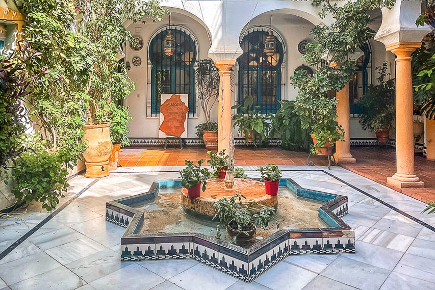 A star-shaped, tiled fountain sits in the middle of a small patio, with marble flooring around it, and white arches on tan columns surounding the courtyard, potted plants all around, and arched blue windows in the walls behind.