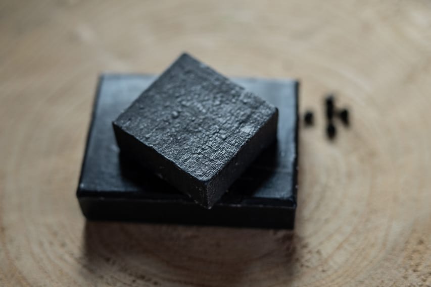 Two squares of black charcoal soap are stacked on each other, sitting on a wooden slab. Black soap is used in traditional Moroccan hammans.