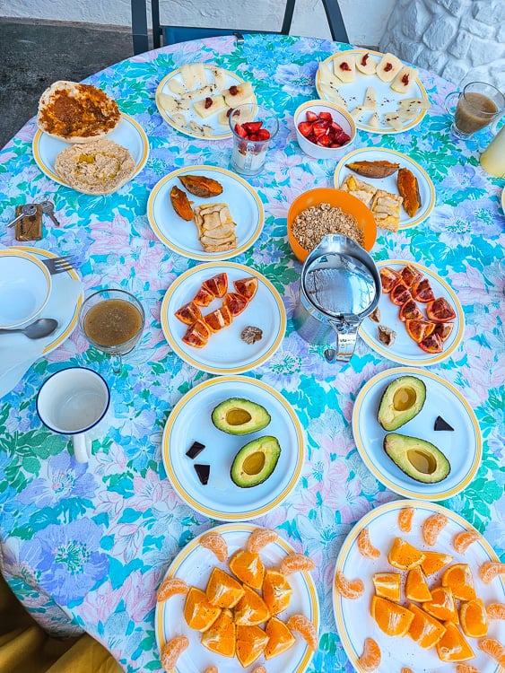 A table set with 10+ plates with different fresh fruits, avocados, breads, and granola, in a white village in Spain.