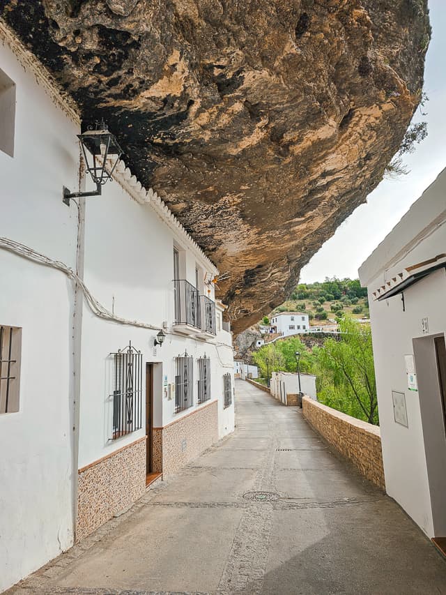 A large rock cliff overhangs a street with white buildings in Setenil. The street is Calle Cabrerizas.