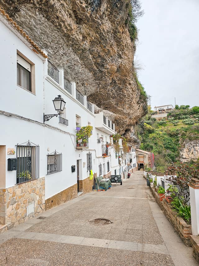 On Calle Calanas, the white buildings sport numerous flower pots and the green hillside is in the background, adding color to contrast the tan cliff.
