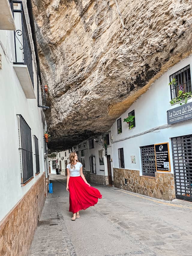 A woman in a red skirt walks down a street in Setenil, Spain. White buildings meld into a slanted, massive rock that covers the street.