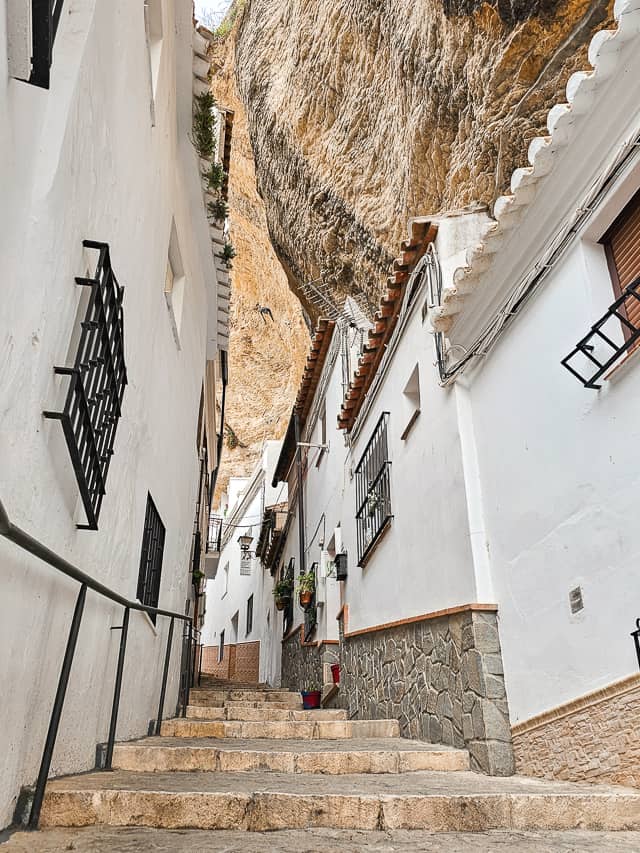 Several steps lead up an alley between white washed buildings with massive cliffs rising out of the buildings. Calle Herreria.