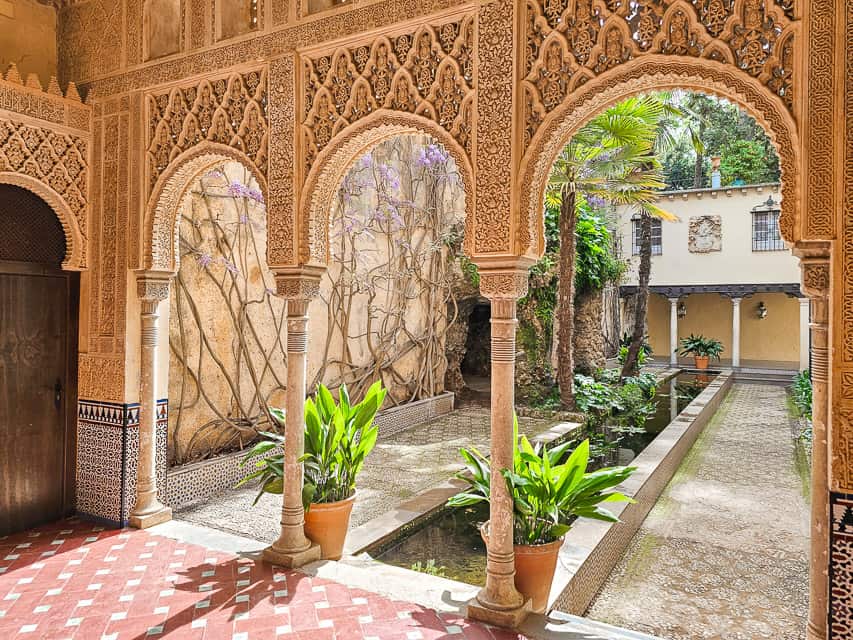 A small garden area with arches in the foreground, a long basin of water strecthing along the garden, and plants, trees, and wisteria in the background.