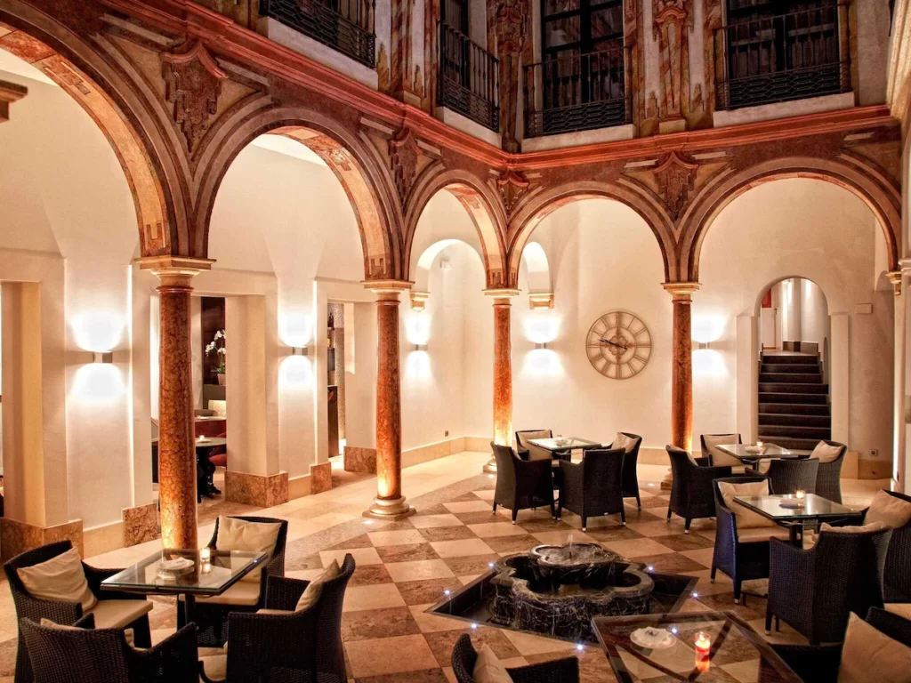 A hotel patio witih carved arches, white walls, checkerboard tiled flooring, and little tables and chairs set up. It's night, and the room is lit cozily.