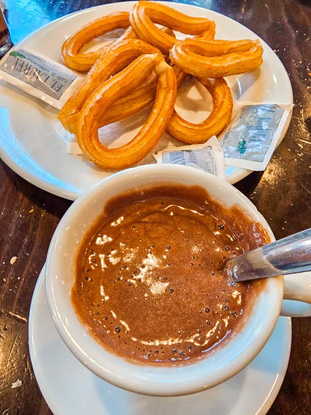 A plate full of curved, skinny curves, with packet of sugar next to them, are on a white plate. A cup of thick hot cocolate is next to the churros.